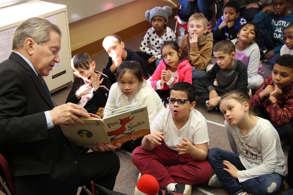 A group of young children sit on the floor watching a man in suit read Fox in Sox