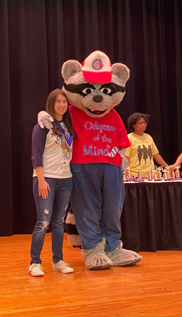 A young woman smiles next to a large costumed racoon with a red shirt and a baseball cap.