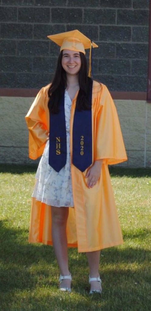 A young woman with long brown hair smiles wearing a gold graduation cap and gown.