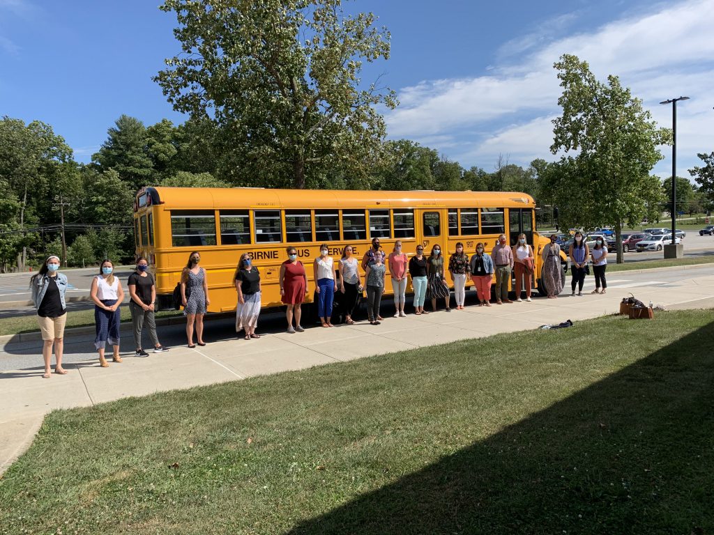 Twenty  teachers wearing masks stand in front of a yellow school bus