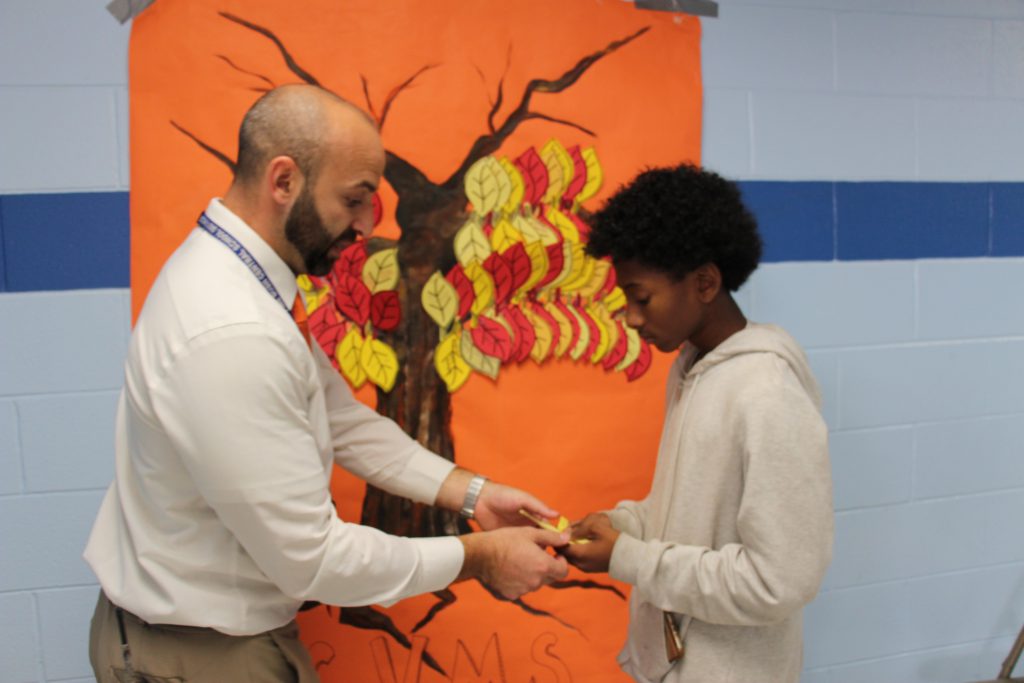 A man with a beard and a middle school student stand in front of an orange background with a tree on it.