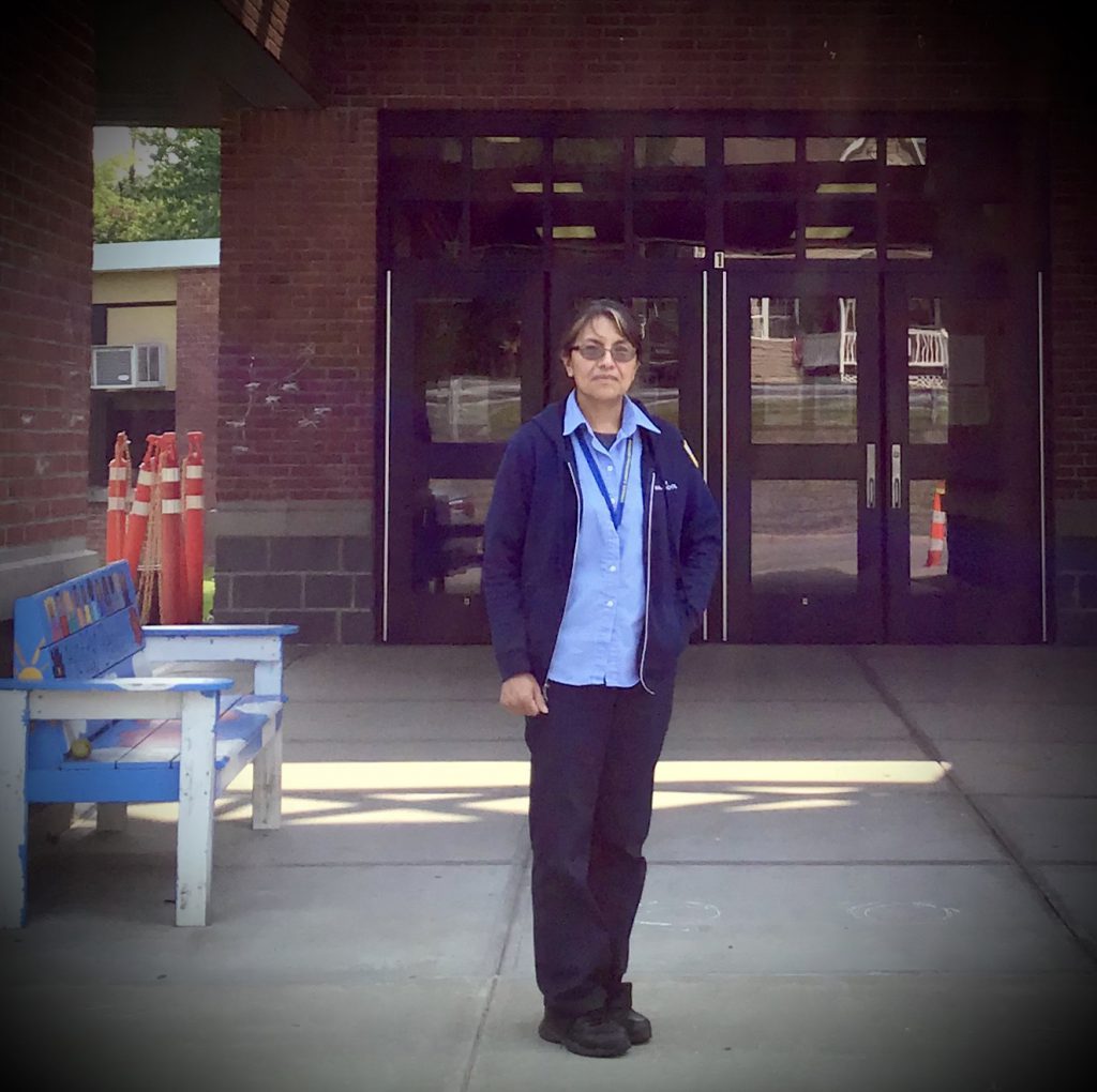 A woman wearing glasses, black pants, a light blue shirt and dark blue jacket stands in front of a brick building