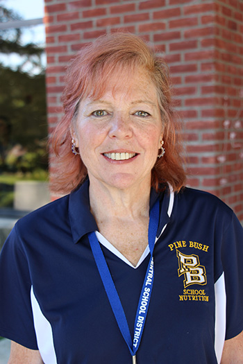 A woman with red hair wearing a blue Pine Bush shirt smiles with a brick building in the background