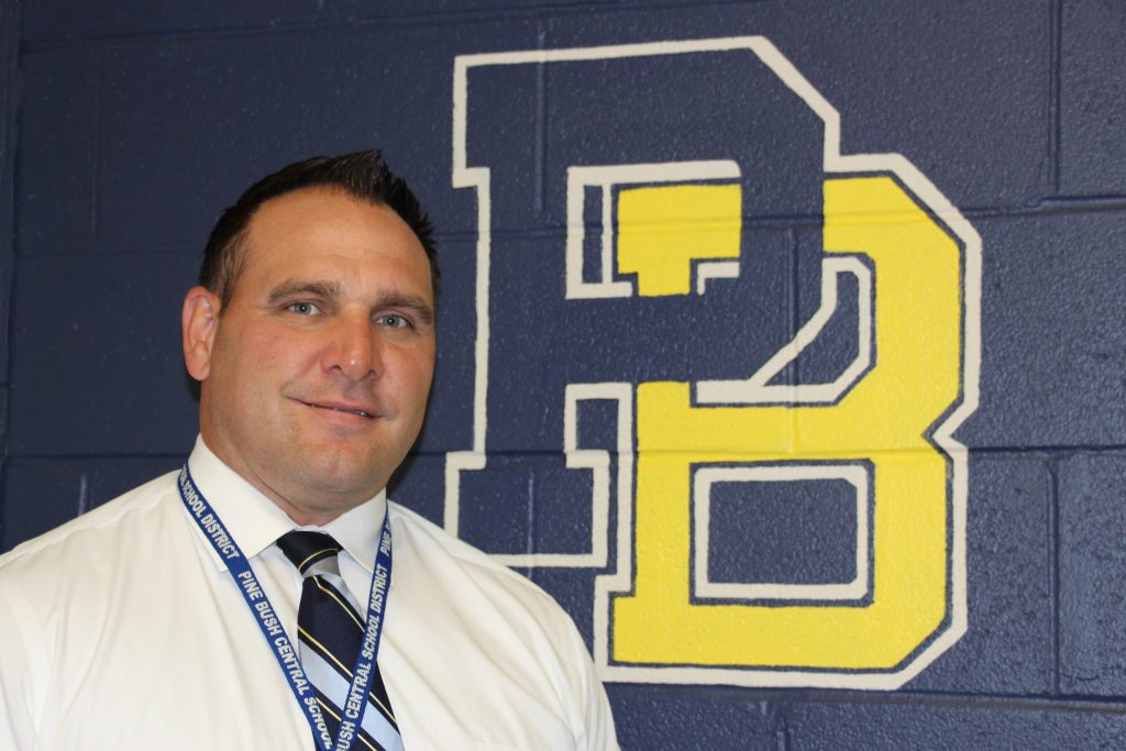 A man with dark hair, wearing a white shirt and blue tie smiles. The blue and gold PB logo is in the background