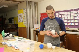 A man with very short hair and beard, wearing a gray shirt with blue sleeves that says PB ARTS holds an object made on a 3-D printer