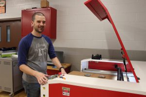 A man with very short hair and beard stands at a large red and white machine.