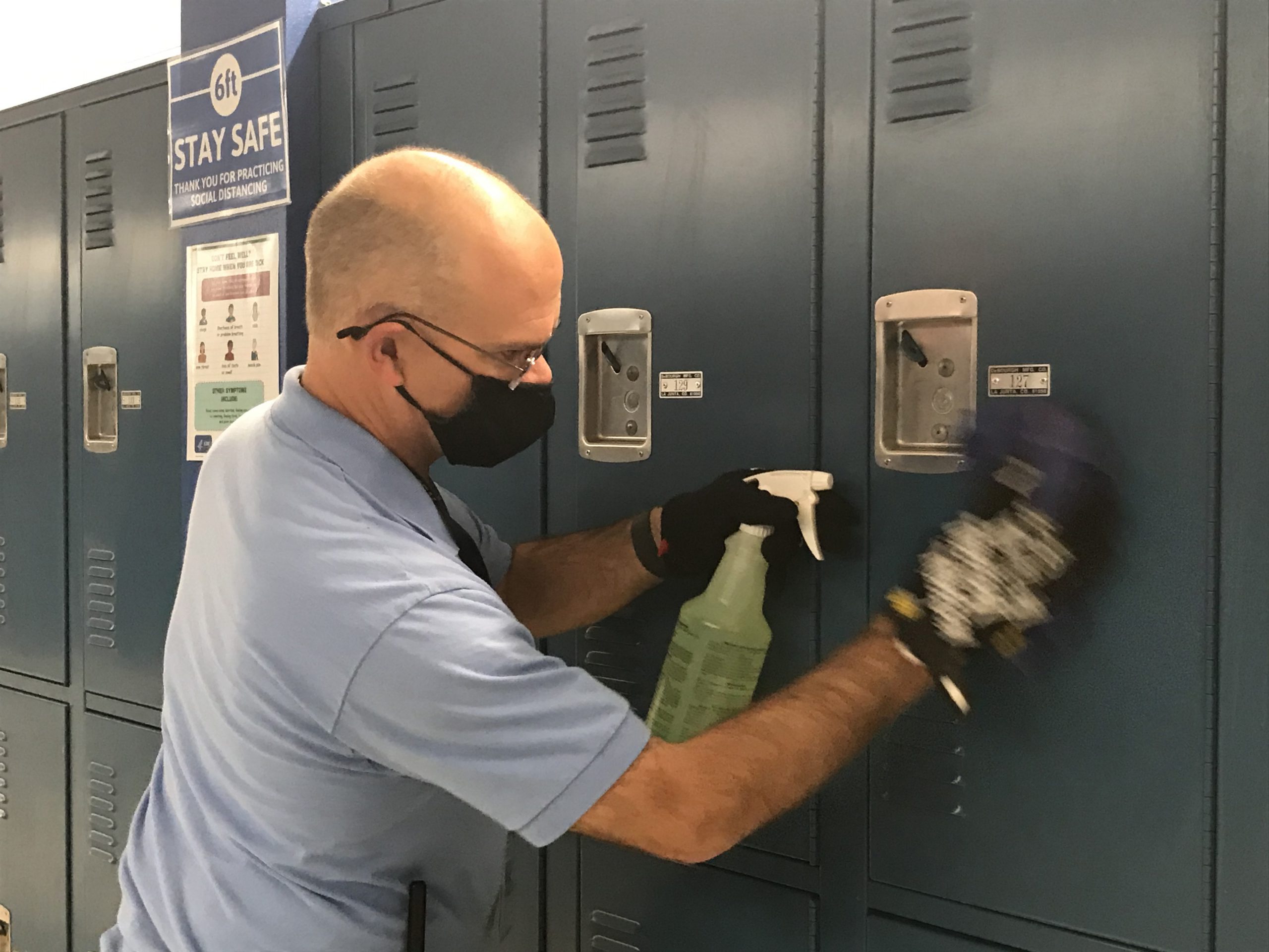 A man wearing a mask and a blue shirt cleans a locker with a cloth and spray bottle