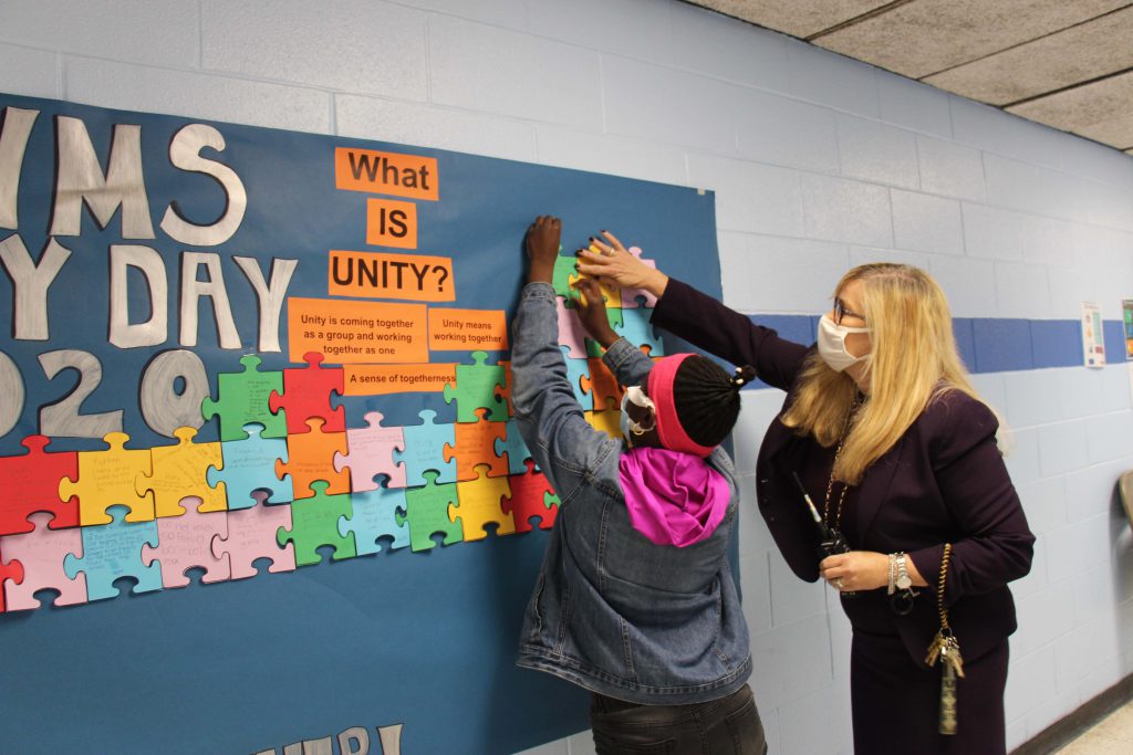 A woman with long blonde hair helps a student wearing a pink and gray sweatshirt  put a paper puzzle piece on a bulletin board