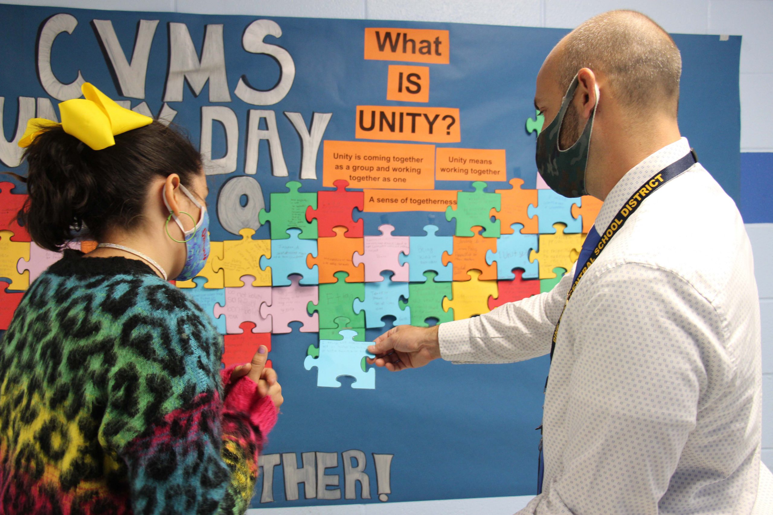 A man wearing a mask holds a blue puzzle piece out in front as a student , also wearing a mask, discusses what she wrote on it