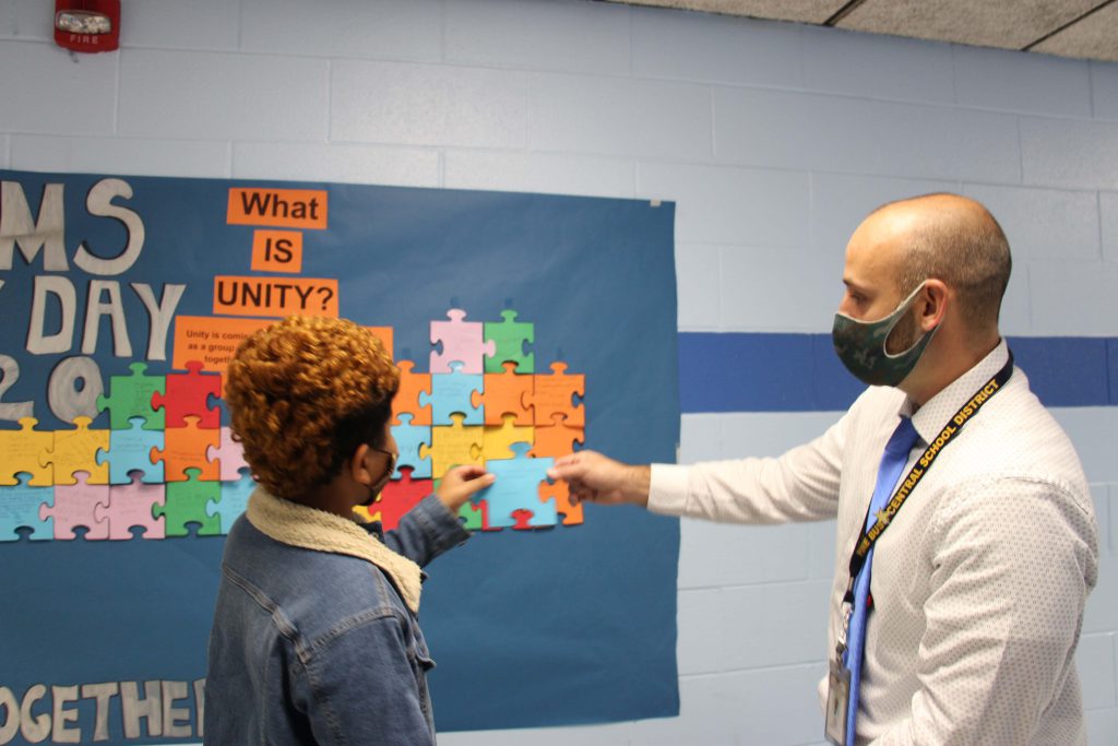 A man holds a paper puzzle piece out while a student wearing a denim jacket reads it