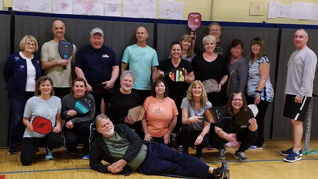 A group of 18 adults stand and kneel holding their pickleball paddles.