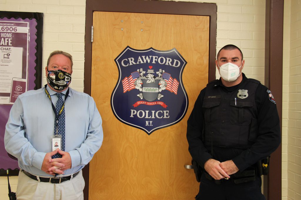 A man in a blue shirt and tie stands on the left side of a door that has a Crawford Police Department shield on it. On the right is a police officer in uniform. Both are wearing masks.