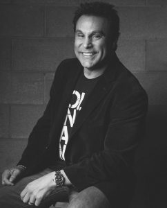 a black and white photo of a man wearing a dark blazer. He has short hair and is smiling.