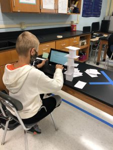 A middle school boy sits at a desk with a Chromebook in front of him, building a tower out of index cards.