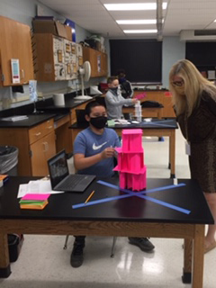 A young man sitting at his desk shows his tower, made of hot pink index cards, to a woman who is standing and watching.