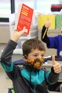 A boy holds up a red dictionary and gives a thumbs up. He is wearing a blue and black shirt and mask.