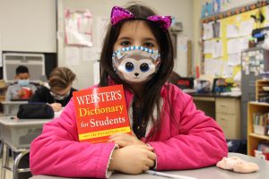 A girl in a pink shirt wearing pink cat ears and a mask sits holding her red dictionary
