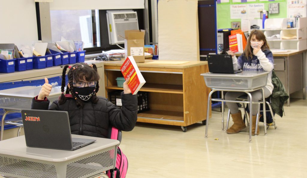 Two girls sitting far apart hold up their red dictionaries and give thumbs up