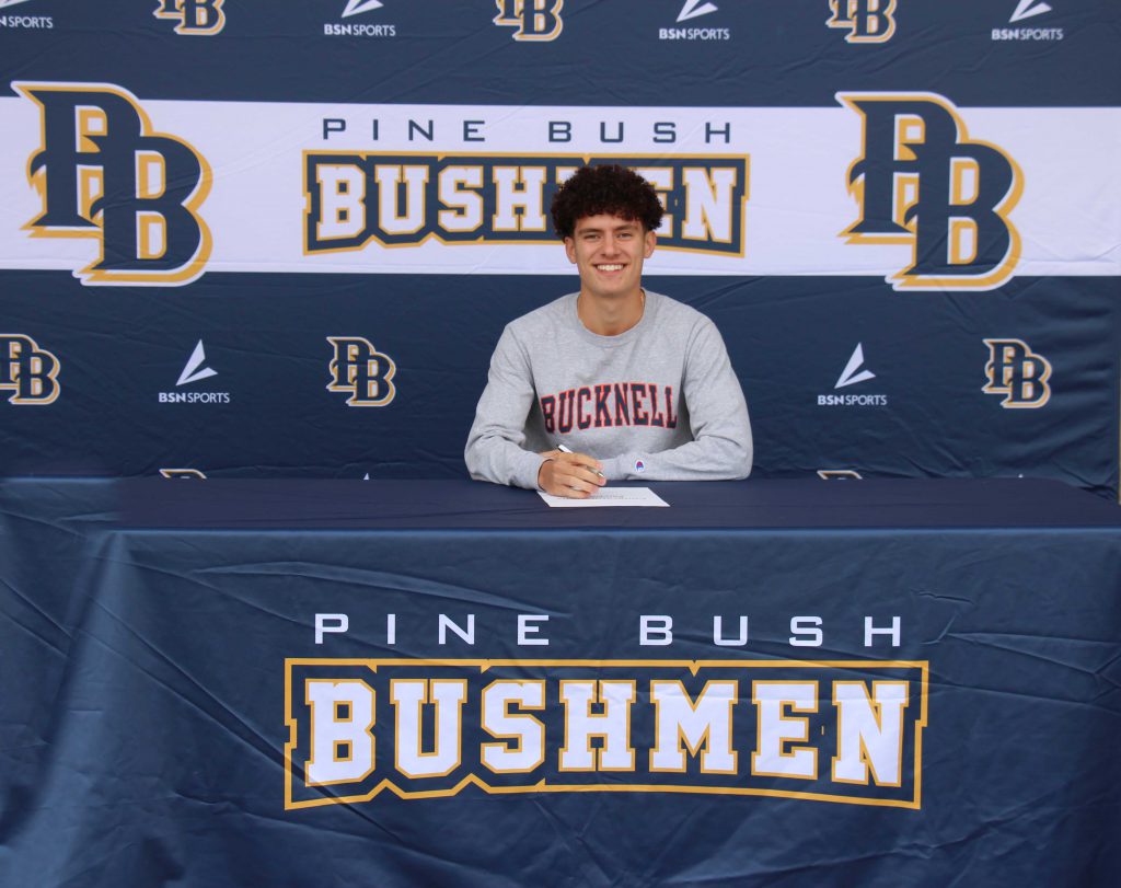 A young man with dark hair and a Bucknell shirt sits at the desk that says Pine Bush Bushmen