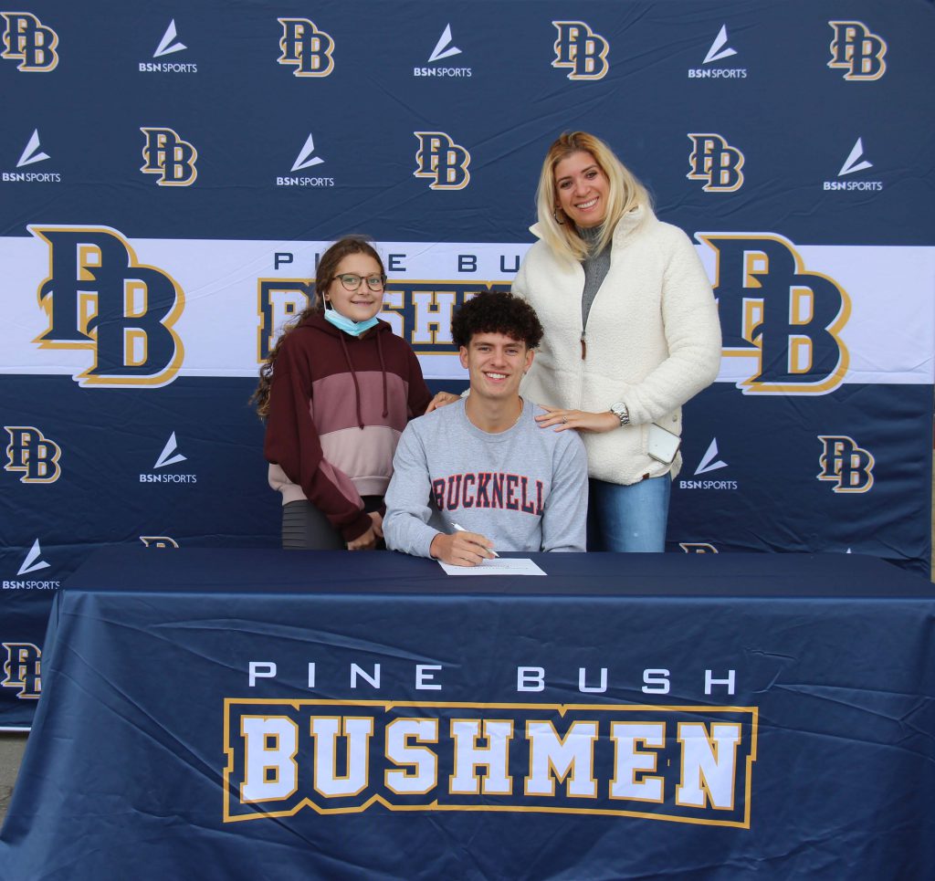 A young girl and a woman stand behind a young man with dark hair wearing a gray shirt that says Bucknell