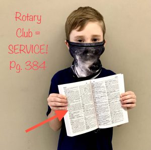 A boy holds his dictionary open to the page that contains the word service. He is wearing a blue and white mask.