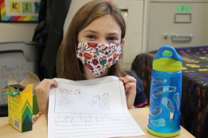 A young girl with brown hair smiles behind a mask and holds up her drawing.