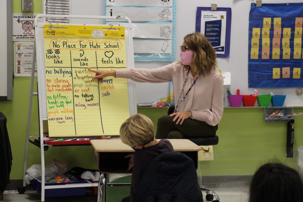 A teacher with a pink mask points to a chart drawn on a large yellow flip chart