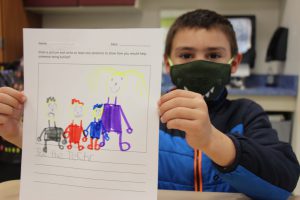 A young boy wearing a black mask with white teeth holds up his drawing which is very colorful