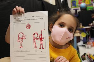 A young student in a yellow shirt holds up a drawing.