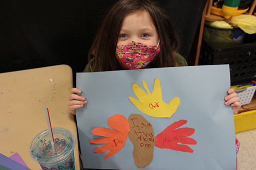 A girl holds up her artwork showing a turkey with handprints listing what she is grateful for.
