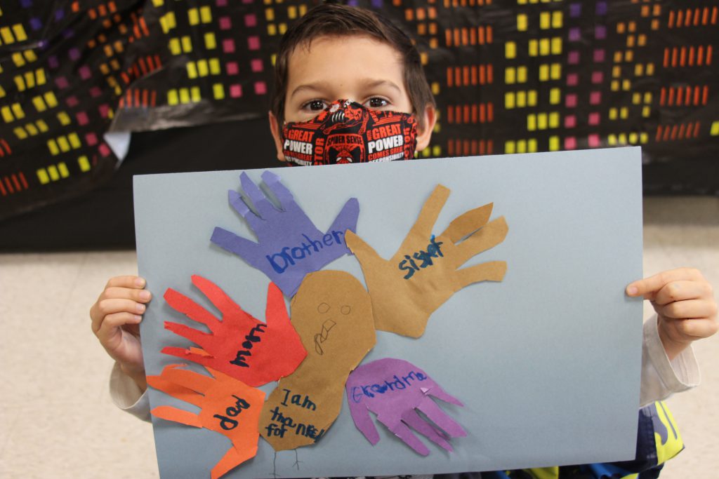 A boy wearing a red, black and white mask holds up his cut-out turkey with handprints saying dad, mom, brother, sister and grandma