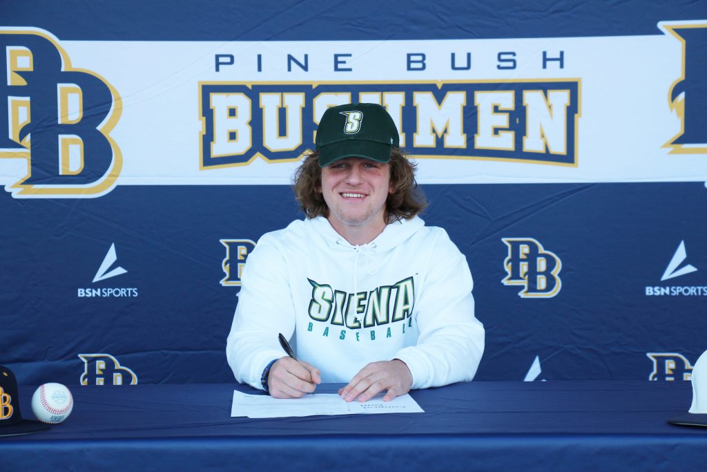 A young man with shoulder-length hair smiles, as he signs a letter. He is wearing a green baseball cap with the letter S on it and a sweatshirt that says Siena. In the background is Pine Bush Bushmen