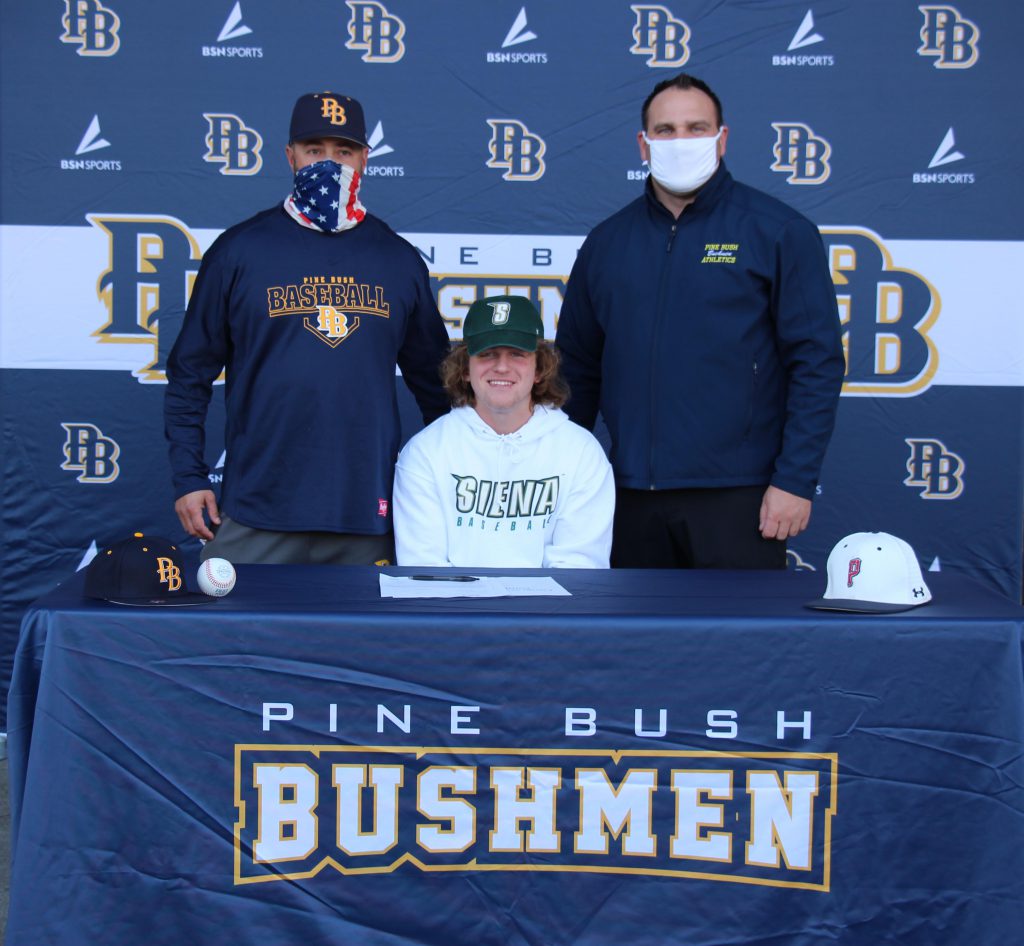 A young man sits at a table with Pine Bush Bushmen on the front and in the background. He is wearing a white Siena sweatshirt. Behind him are two men wearing masks and Pine Bush shirts