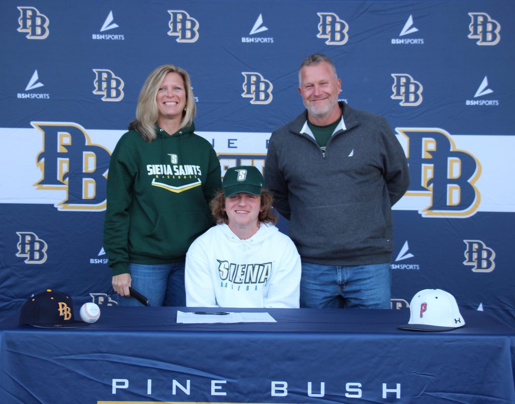 A woman and man stand behind a young man wearing a green baseball cap and white sweatshirt that says Siena. Table and background say Pine Bush Bushmen