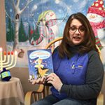 A woman with shoulder-length dark hair wearing a blue and gray sweatshirt that says Pine Bush holds a book. Next to her is a menorah and behind her is a scene with snowmen