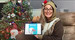 A woman wearing a rudolph hat sits next to a Christmas tree and reads a book