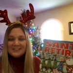 A woman with long blonde hair wearing sparkly red anglers with a Christmas tree in the background holds up a book called Santa Retires