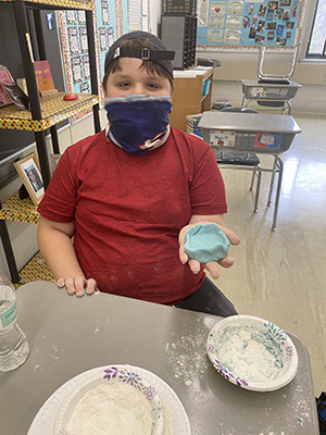 A boy wearing a red shirtand wearing a dark mask holds out his light blue little bowl.