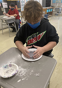 A boy in a black sweatshirt and blue mask is shaping a bowl on his desk.