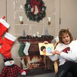 A woman sits in a rocking chair with a fireplace, stockings, wreath and snowmen around her. She is holding a book.