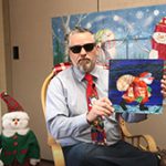A man wearing sunglasses and a funky red tie sits in a rocking chair and holds up a book with a picture of Santa in the snow