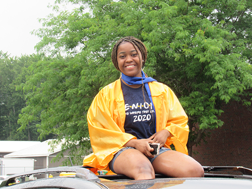 A young woman sits on the top of a car smiling, wearing a gold graduation gown with a blue t-shirt that says Seniors 2020