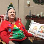 A woman in a red sweater with a large Christmas tree on it and a headband with a Christmas tree sits in a rocking chair reading a children's book