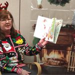 A woman with  festive Christmas sweater and a headband with antlers reads a book in front of a fireplace