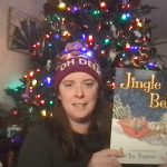 A woman with a knit hat on that says oh Deer holds up a book called Jingle Bells. There is a lit Christmas tree in the background