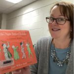 A woman with glasses and a blue sweater and necklace holds up an orange book