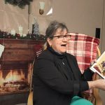 A woman with glasses, wearing a black jacket, sits in front of a fireplace reading a book