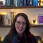 A woman with long brown hair and glasses smiles. She is sitting in front of book shelves