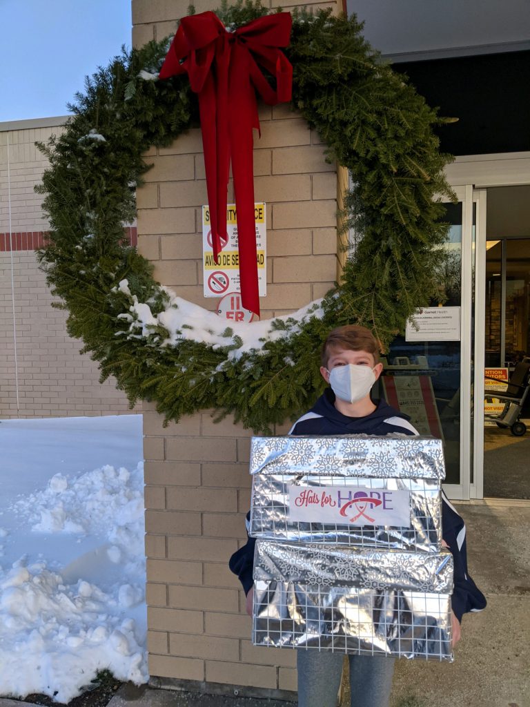 A young man wearing a blue mask holds a box covered in silver paper in front of a large wreath. The box says Hats for Hope.