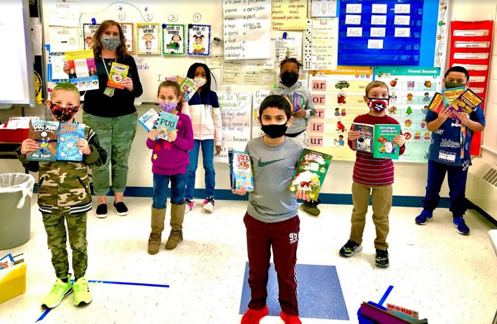 six students and a teacher stand separated and wearing masks holding several books each